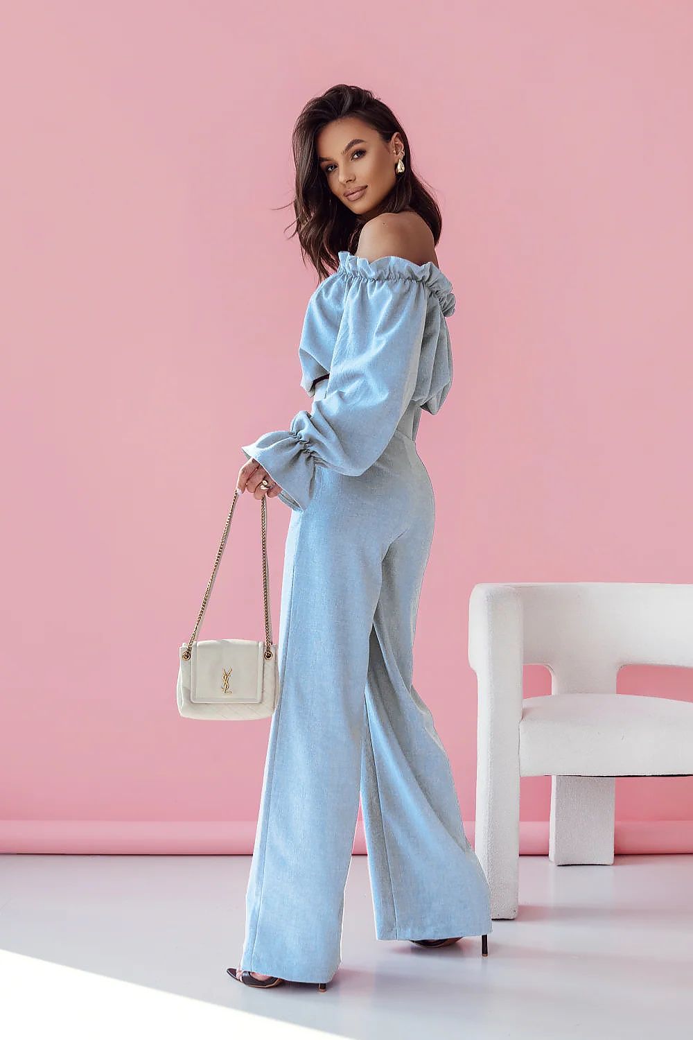 Woman in light blue off-shoulder top and wide-leg pants holding a white handbag against a pink wall.