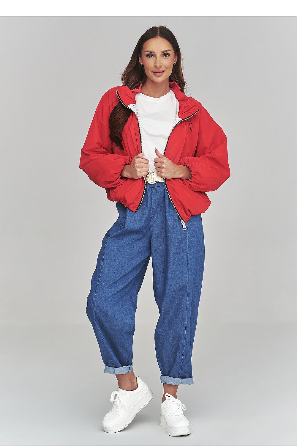 Woman wearing a red jacket, white shirt, blue jeans, and white sneakers on a gray background