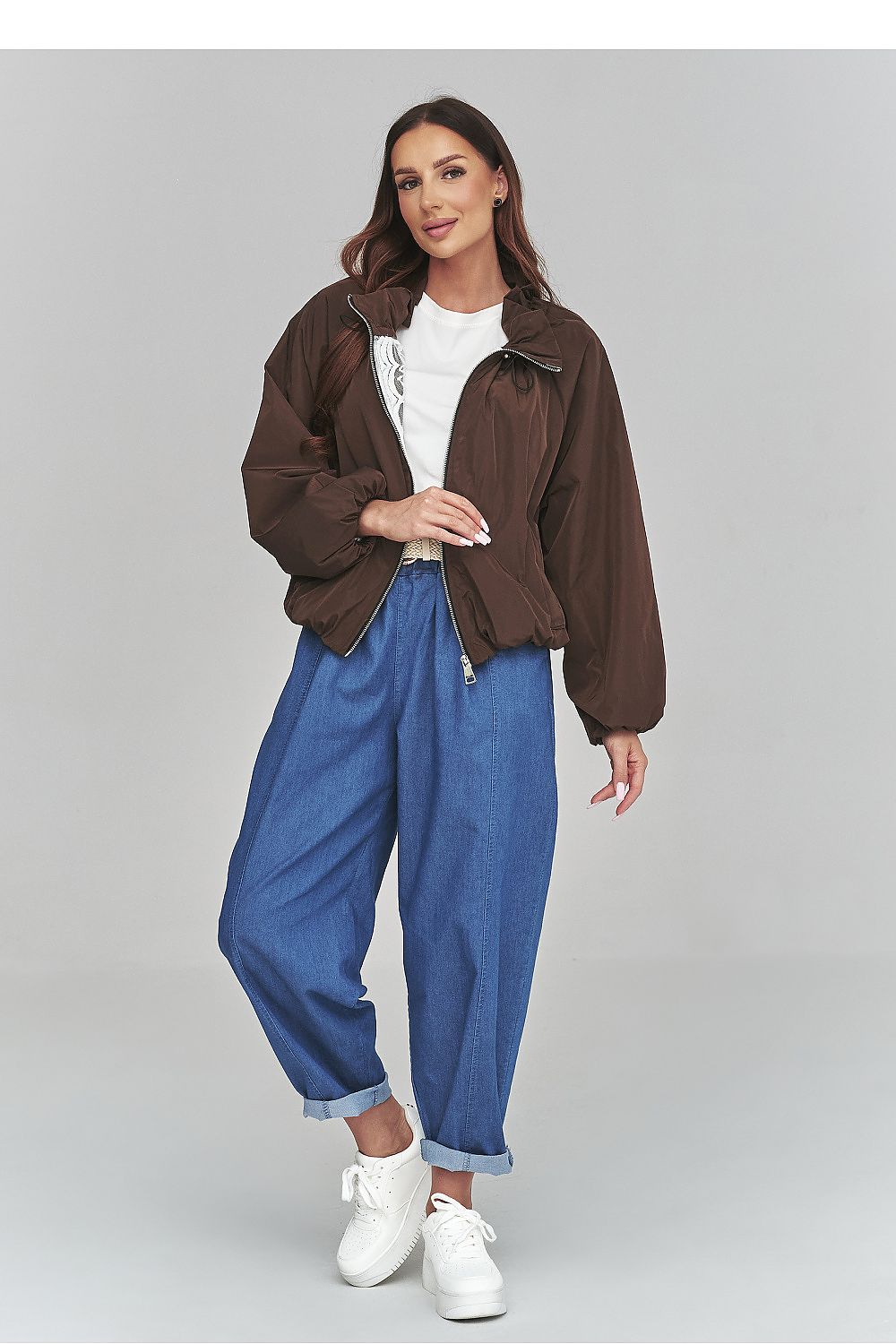Woman wearing a brown jacket, white shirt, blue jeans, and white sneakers on a gray background