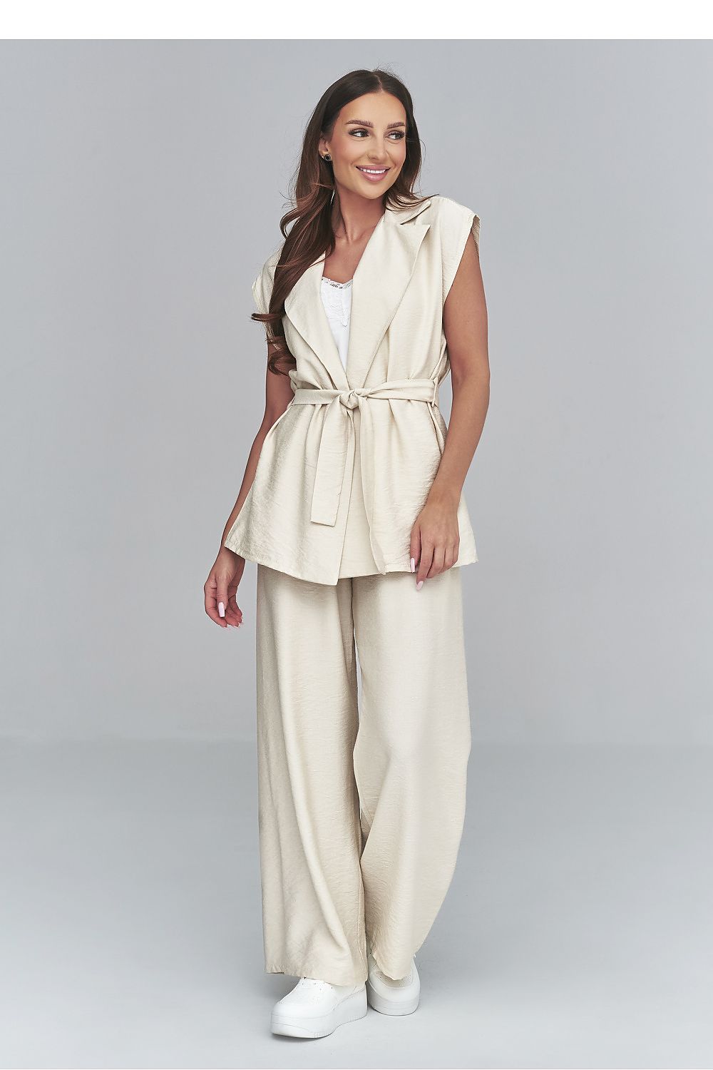 Woman wearing a beige sleeveless jacket and pants set on a gray background