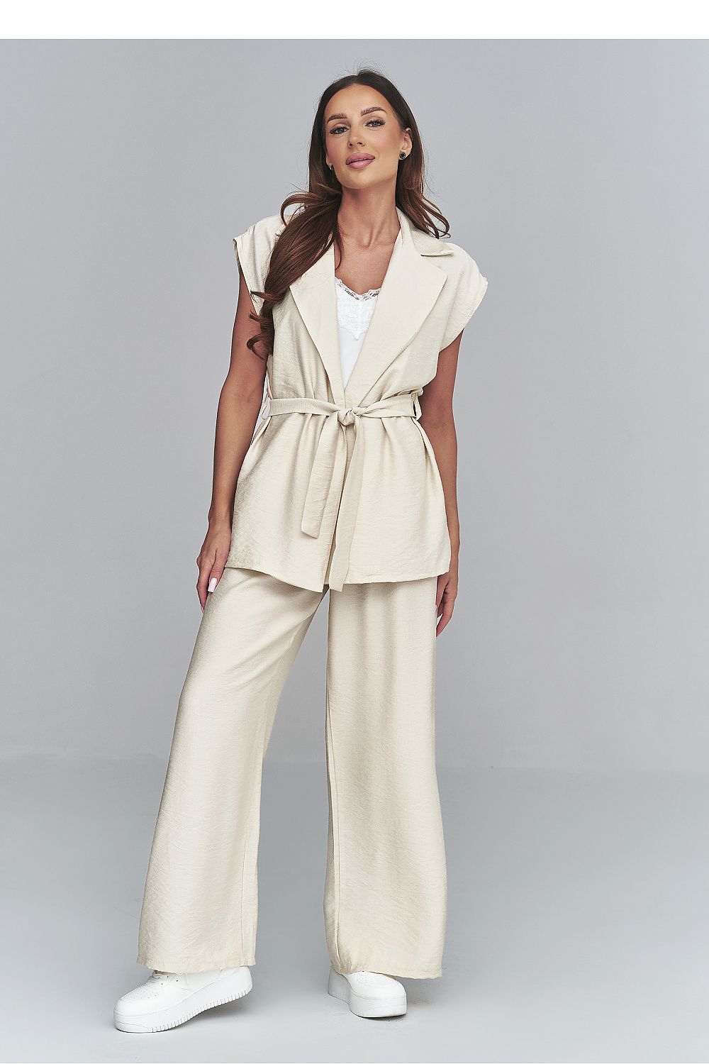 Woman wearing a beige sleeveless jacket and wide-leg pants on a gray background