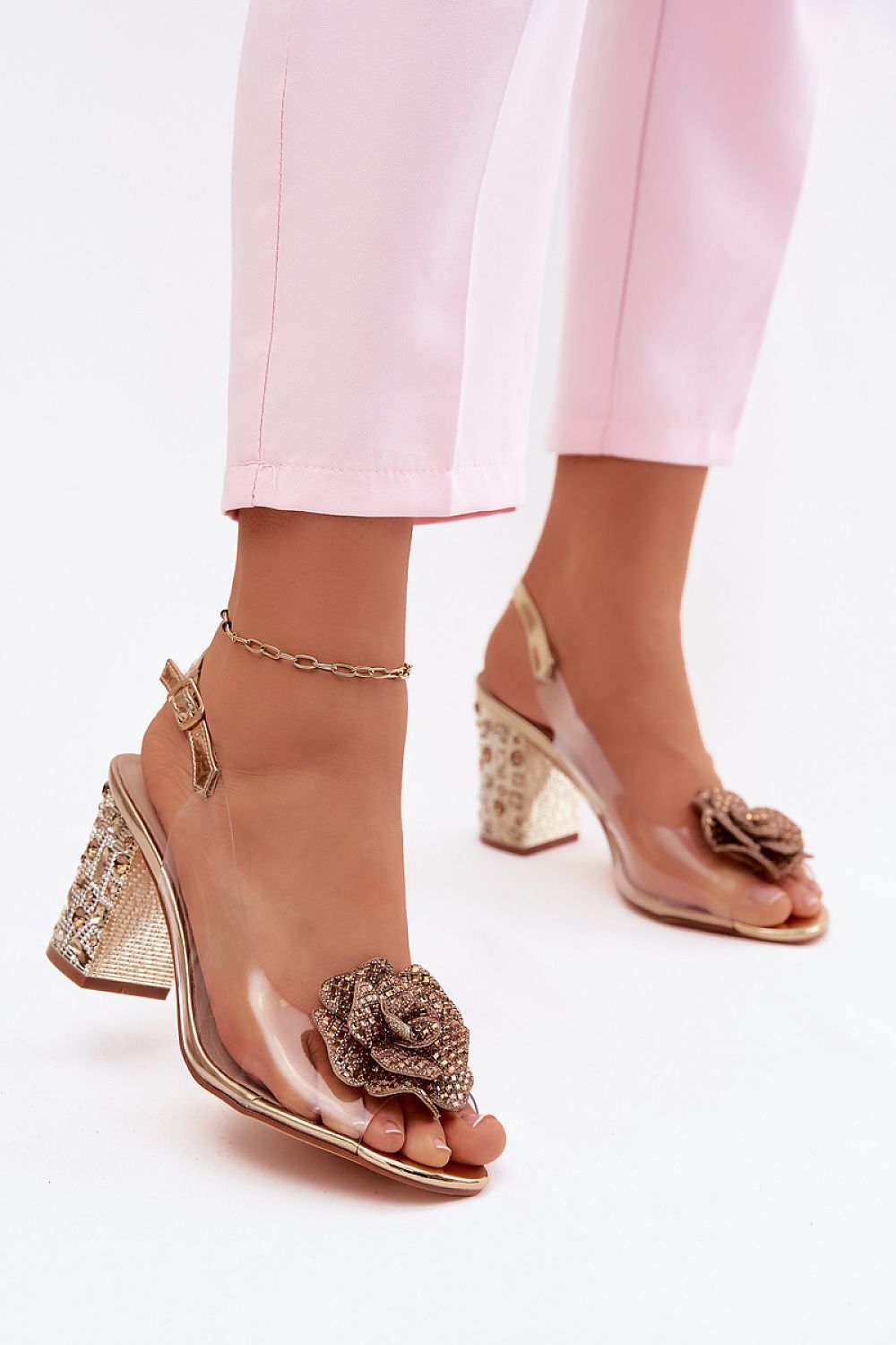 Gold high-heeled shoes with floral embellishments worn with light pink pants on a white background