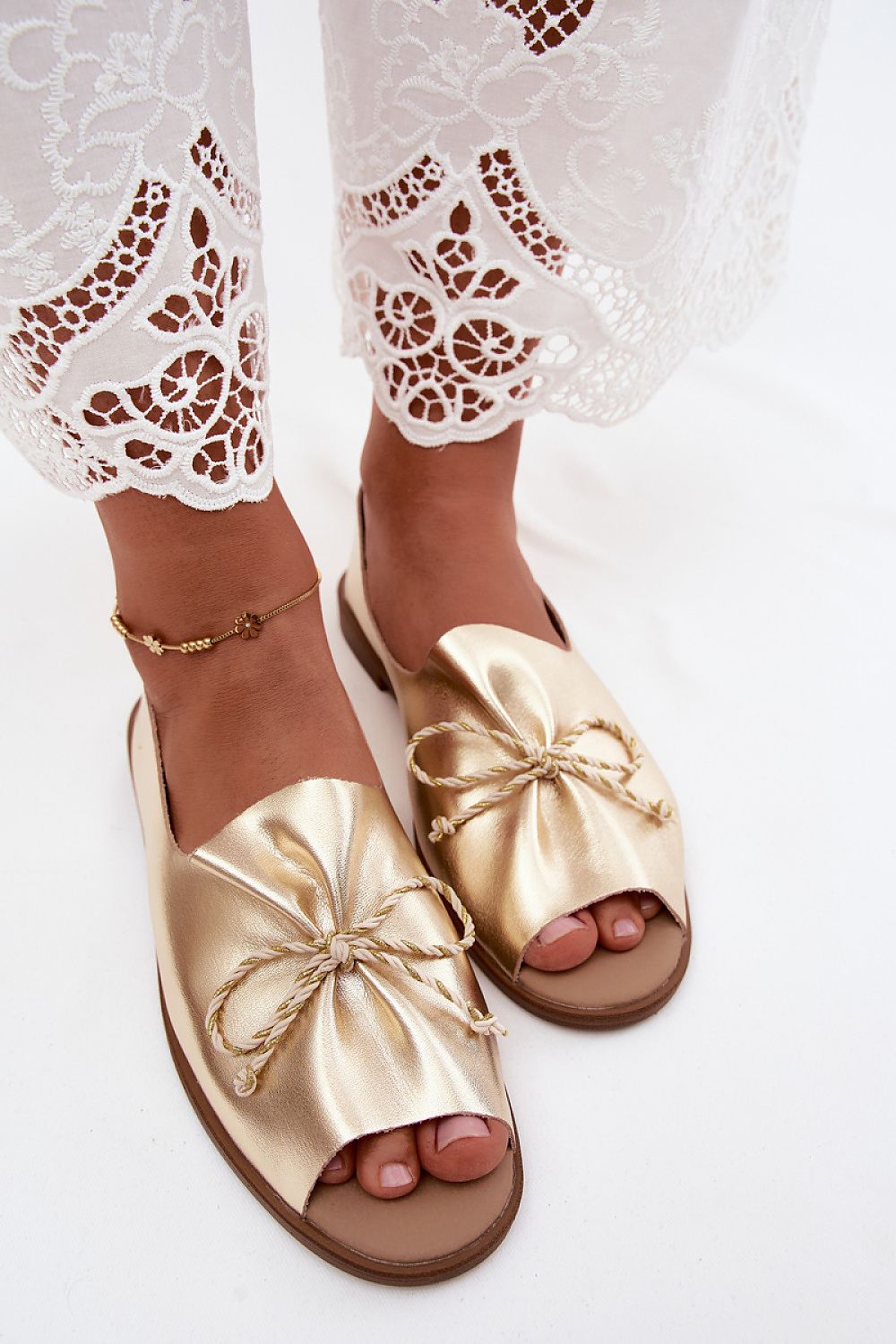 Gold sandals with bow details worn with white lace pants on a light background