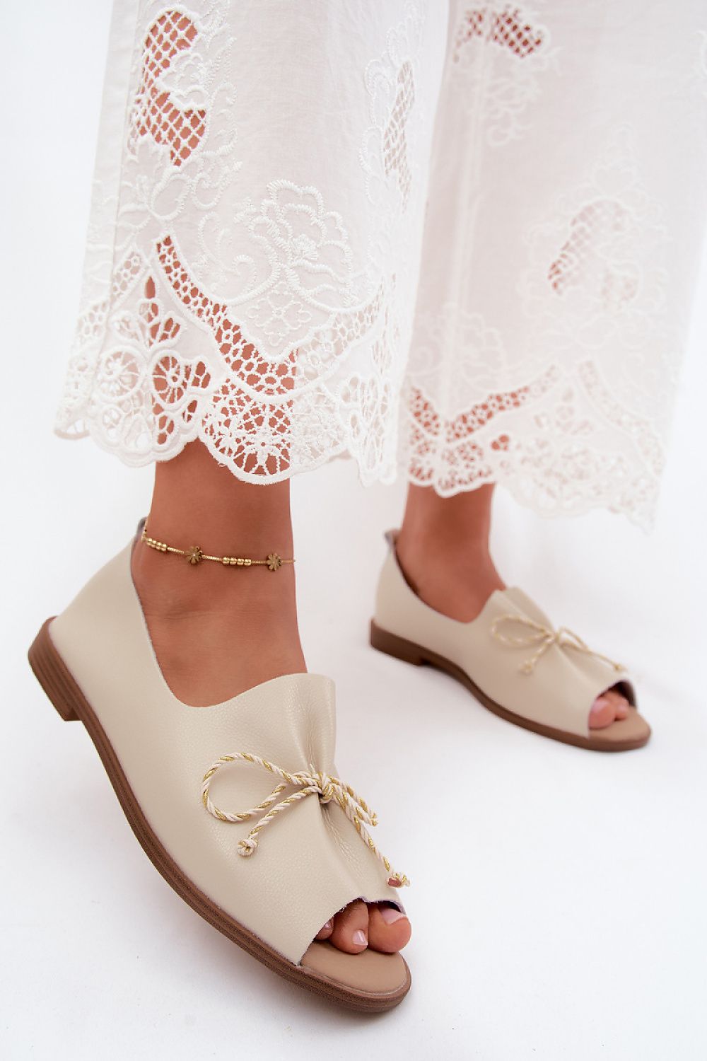 Beige open-toe shoes with a bow detail worn with white lace pants on a white background