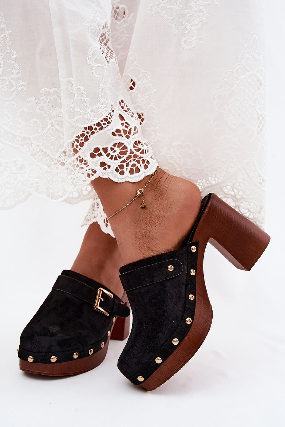 Black suede clogs with brown soles worn by a person on a white lace background