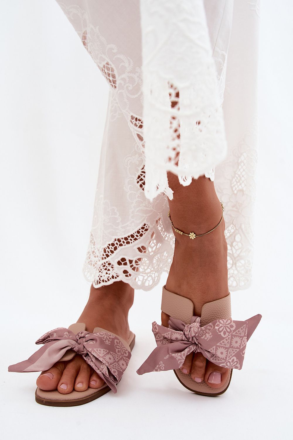 Brown sandals with pink bow details worn with white lace pants on a white background