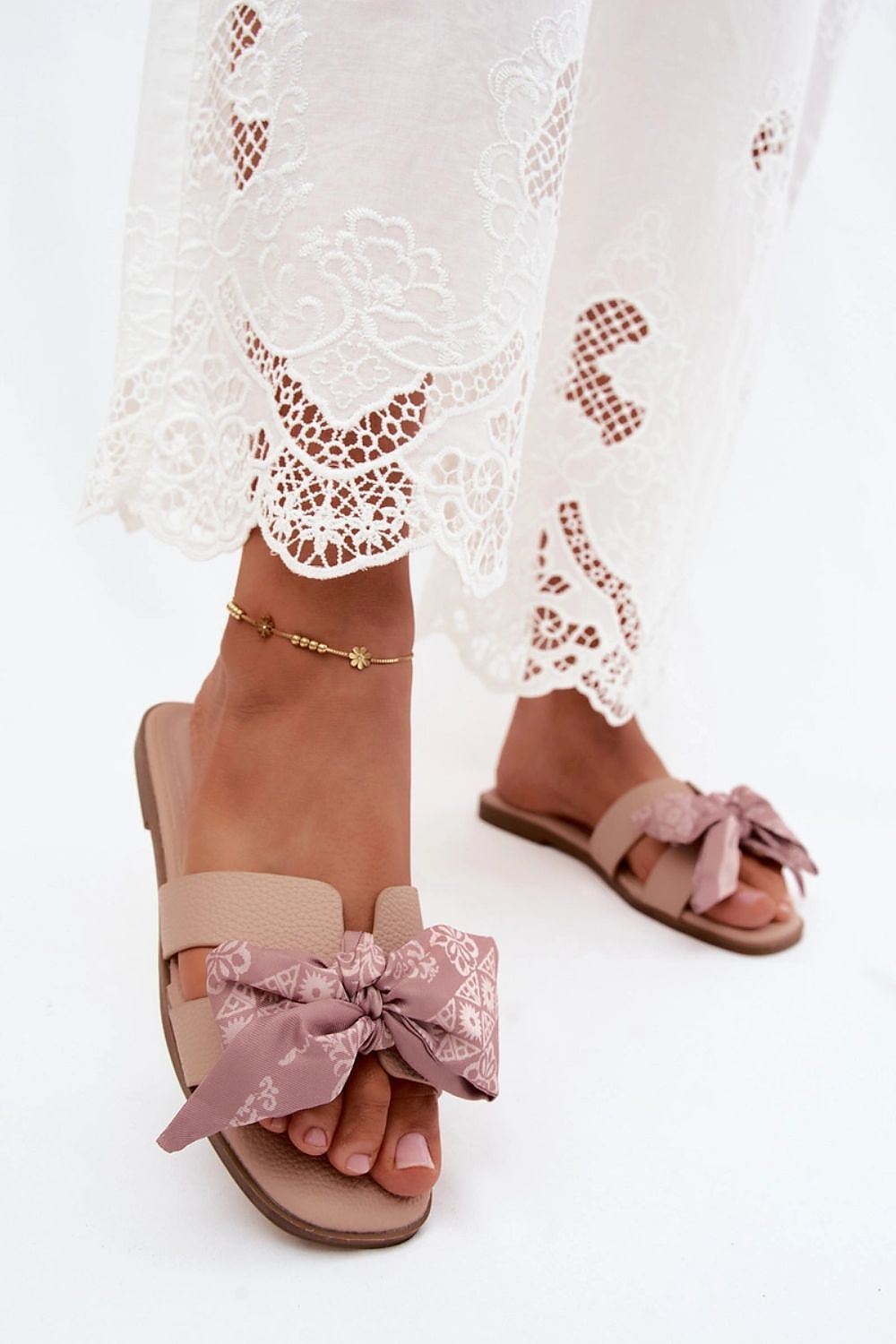 Pink sandals with floral bows worn with a white lace dress on a white background