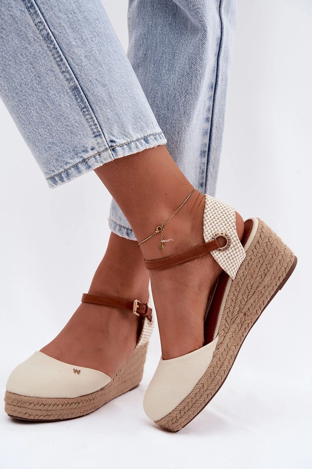 Pair of beige and brown wedge sandals with espadrille sole worn with light blue jeans on a white background.