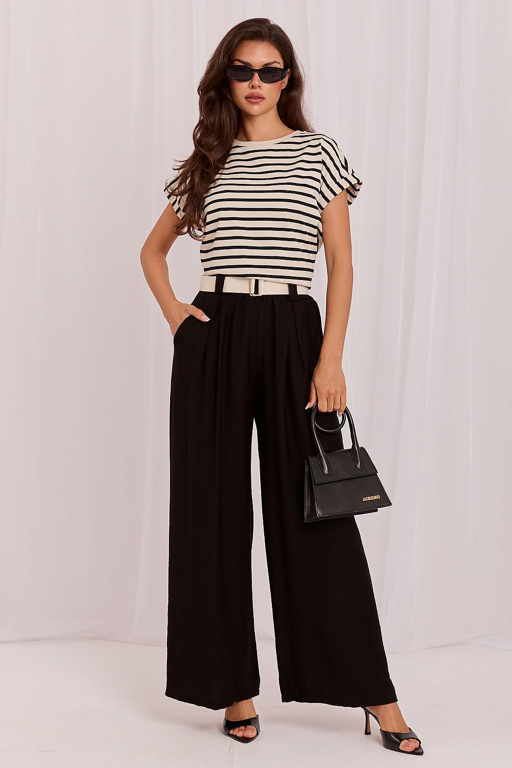 Woman wearing a black and white striped top with wide-leg pants, holding a black handbag on a white background.