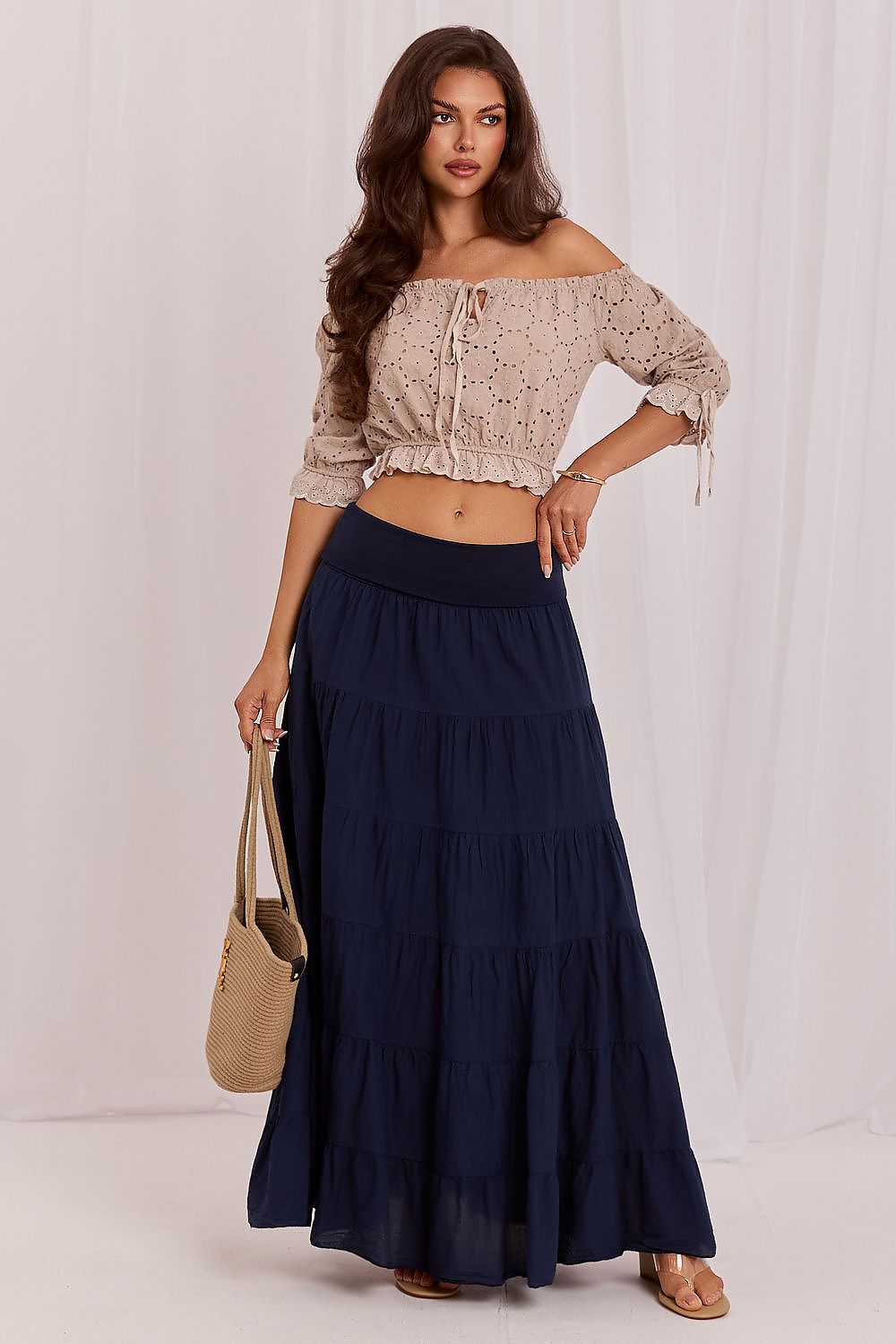 Woman wearing a beige off-shoulder top and navy blue tiered skirt with a beige handbag on a white background