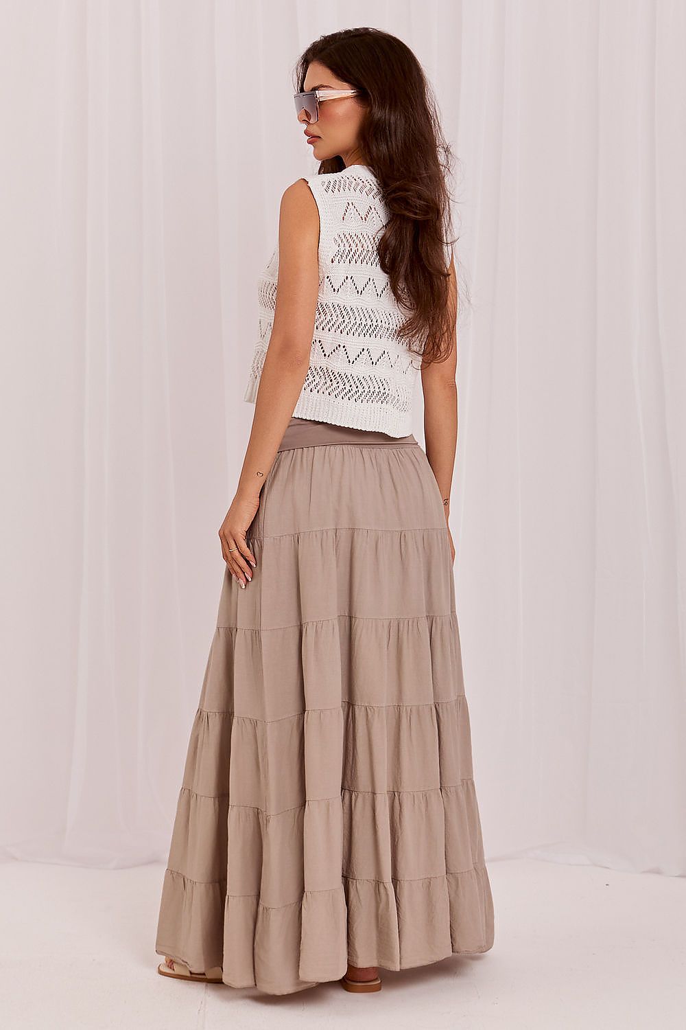 Woman wearing a white sleeveless top with geometric pattern and a long beige tiered skirt on a plain background