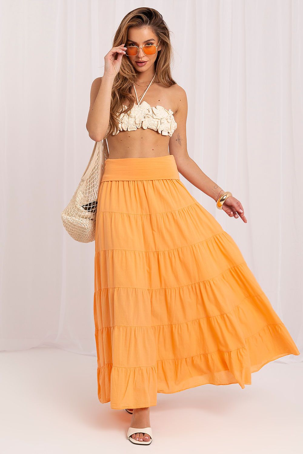 Woman wearing a yellow top and orange skirt with sunglasses and a bag on a white background