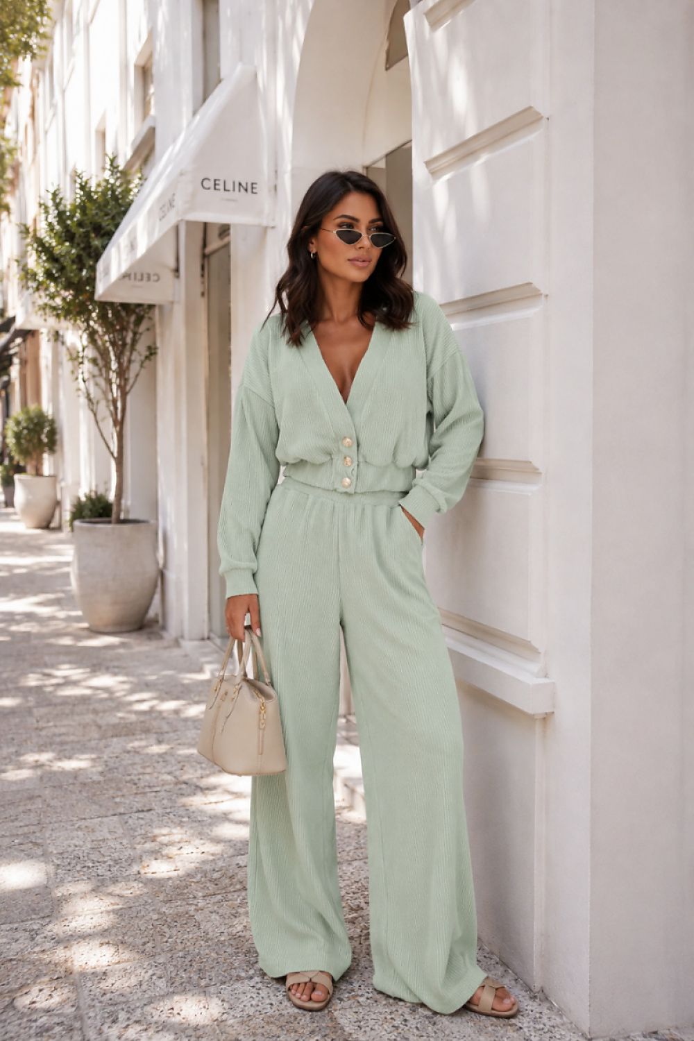 A woman stands on a sunlit street corner wearing a light sage green jumpsuit with a deep V-neck, long sleeves, and wide legs. She is holding a beige handbag and wearing sunglasses.