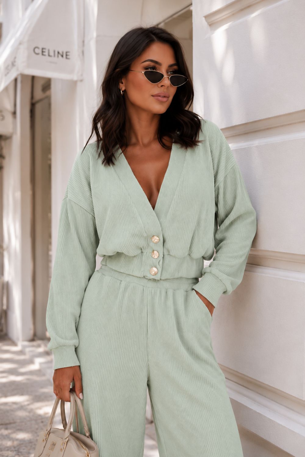 A woman wearing a light sage green ribbed jumpsuit with a deep V-neck, button-front waist, and wide legs, standing outdoors against a white building wall. She is also wearing sunglasses and holding a small handbag.