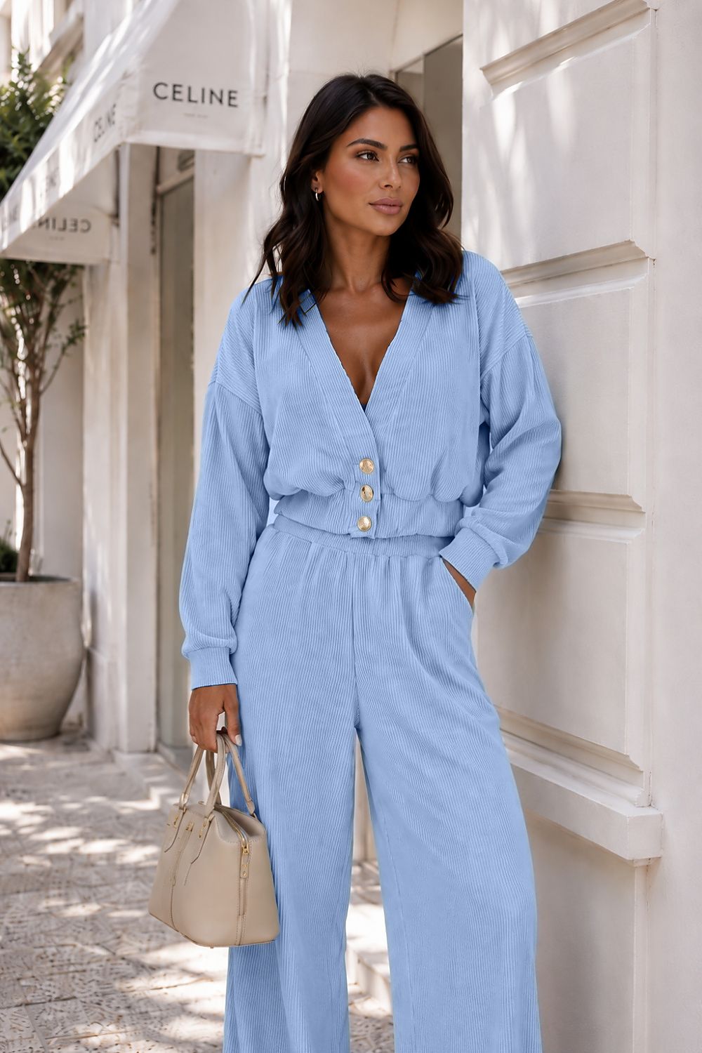A woman wearing a light blue long-sleeve jumpsuit with a buttoned V-neck bodice and wide legs, standing outdoors against a white building wall and holding a beige handbag.