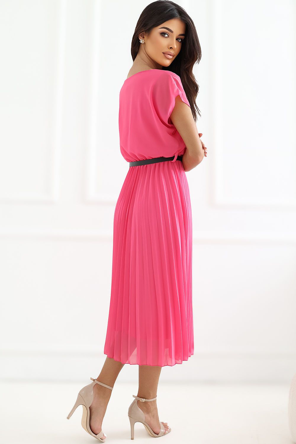 A woman modeling a bright pink midi dress with a short cap-sleeve, pleated skirt, and a thin black waist belt, photographed against a clean white studio background.