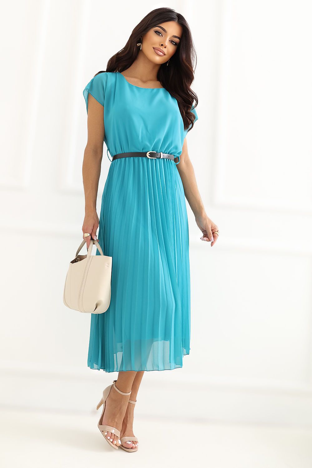 A woman modeling a turquoise blue midi dress with a thin black belt, holding a cream-colored handbag and wearing beige high-heeled sandals against a white studio background.