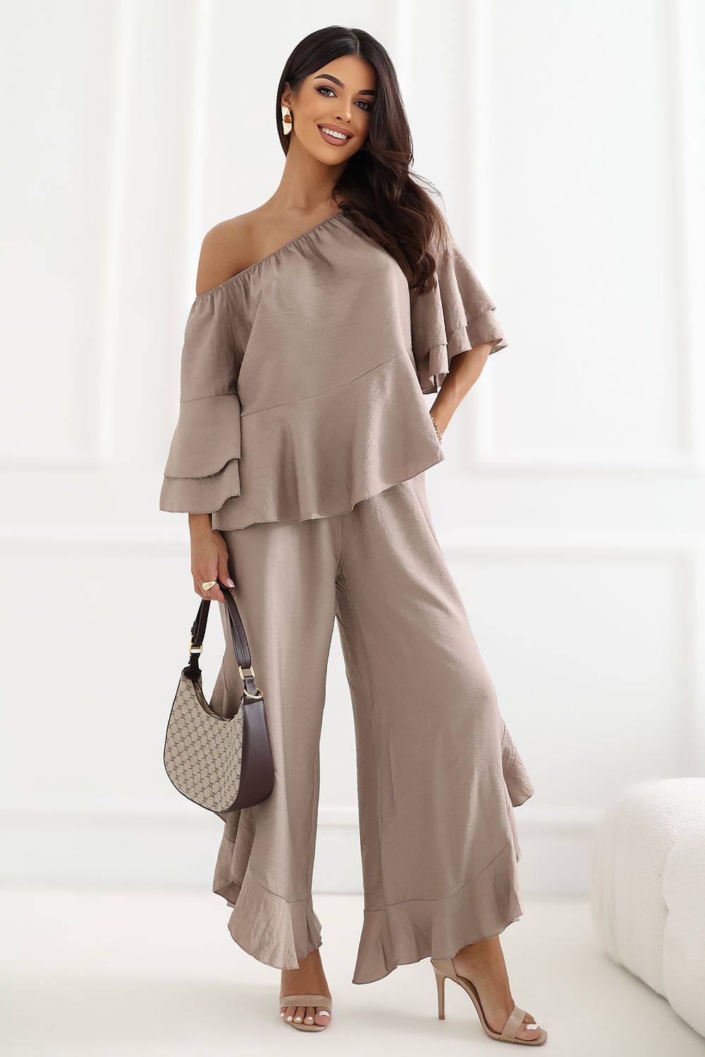A woman models a taupe-beige two-piece outfit featuring an off-the-shoulder ruffled top and matching wide-leg ruffled trousers, styled with a patterned handbag and nude heels.