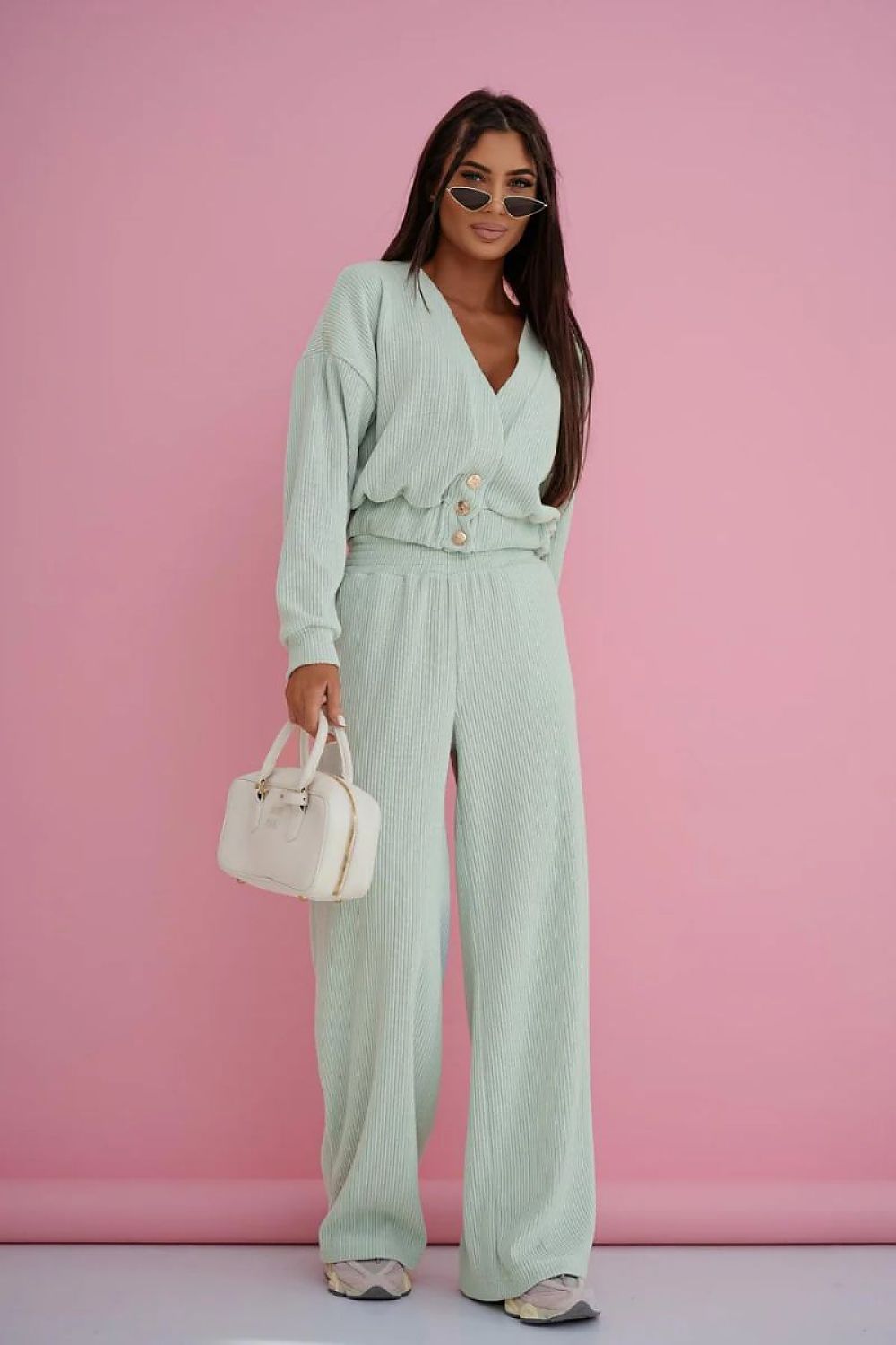A woman modeling a mint green two-piece lounge set consisting of a long-sleeve button-front top and matching wide-leg trousers, holding a small white handbag against a solid pink studio backdrop.