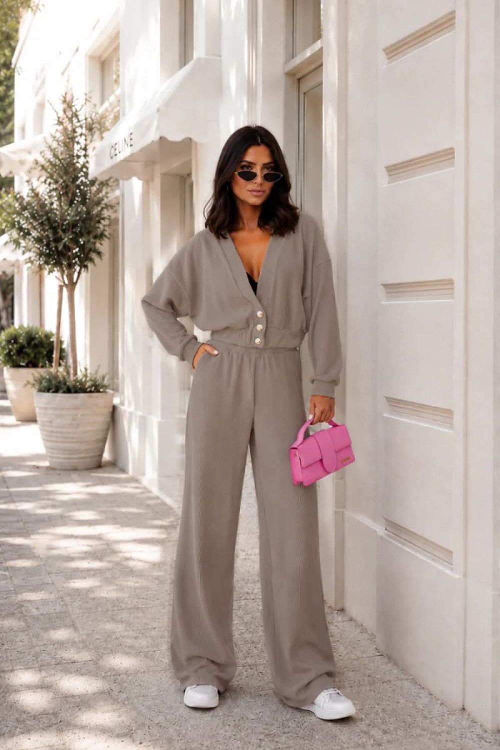 A woman stands on a sunlit sidewalk beside a white building, wearing a taupe-gray wide-leg jumpsuit with a button-front waist, white sneakers, and holding a bright pink handbag.