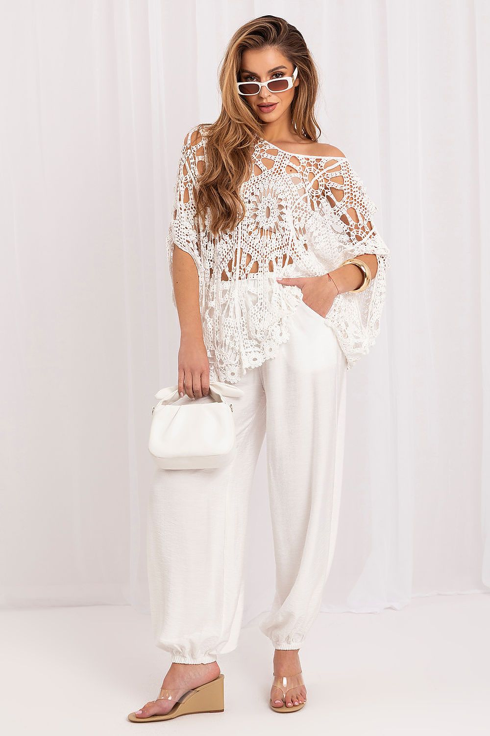 A woman modeling a white crochet lace off-the-shoulder top with matching white wide-leg pants, holding a small white handbag and wearing clear-strap wedge sandals against a white draped backdrop.