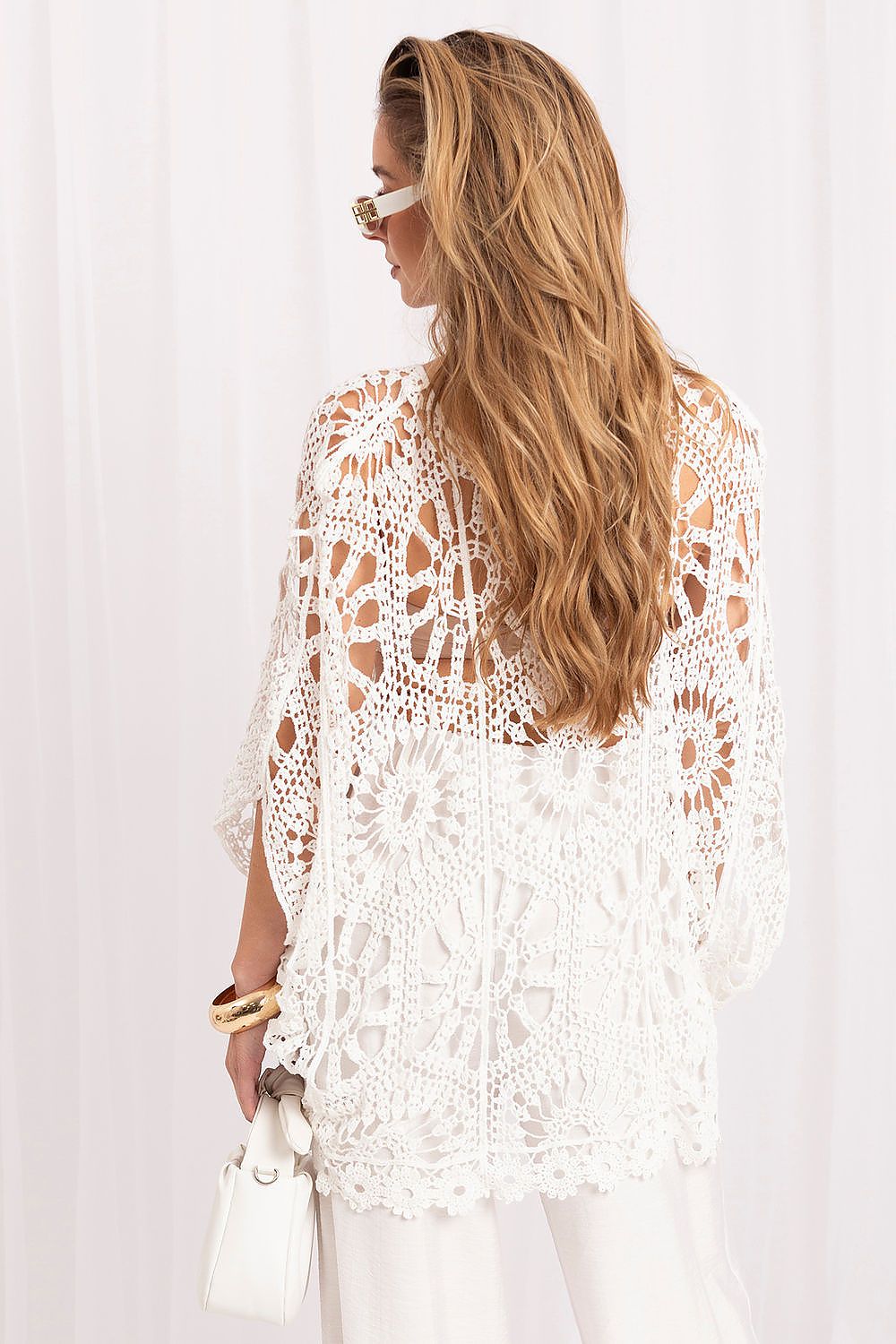 A woman shown from the back wearing a white crochet lace top with sheer sleeves and a scalloped hem, paired with white pants and holding a small white handbag against a soft white curtain background.