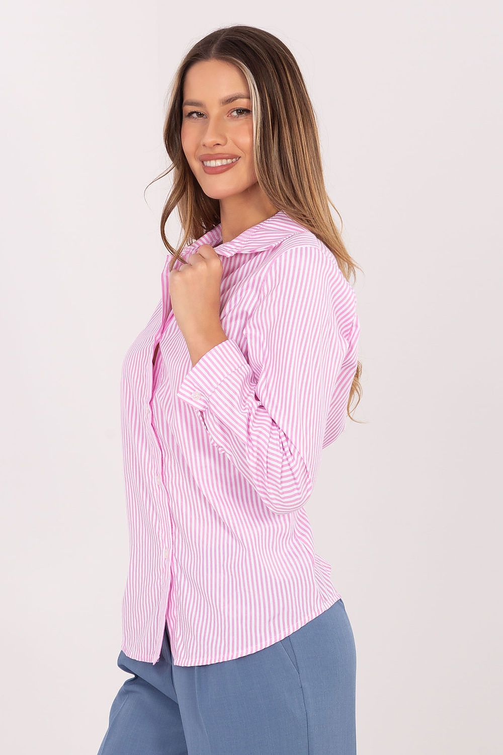 A woman wearing a pink and white vertically striped button-down shirt with blue pants, posing against a light off-white studio background.