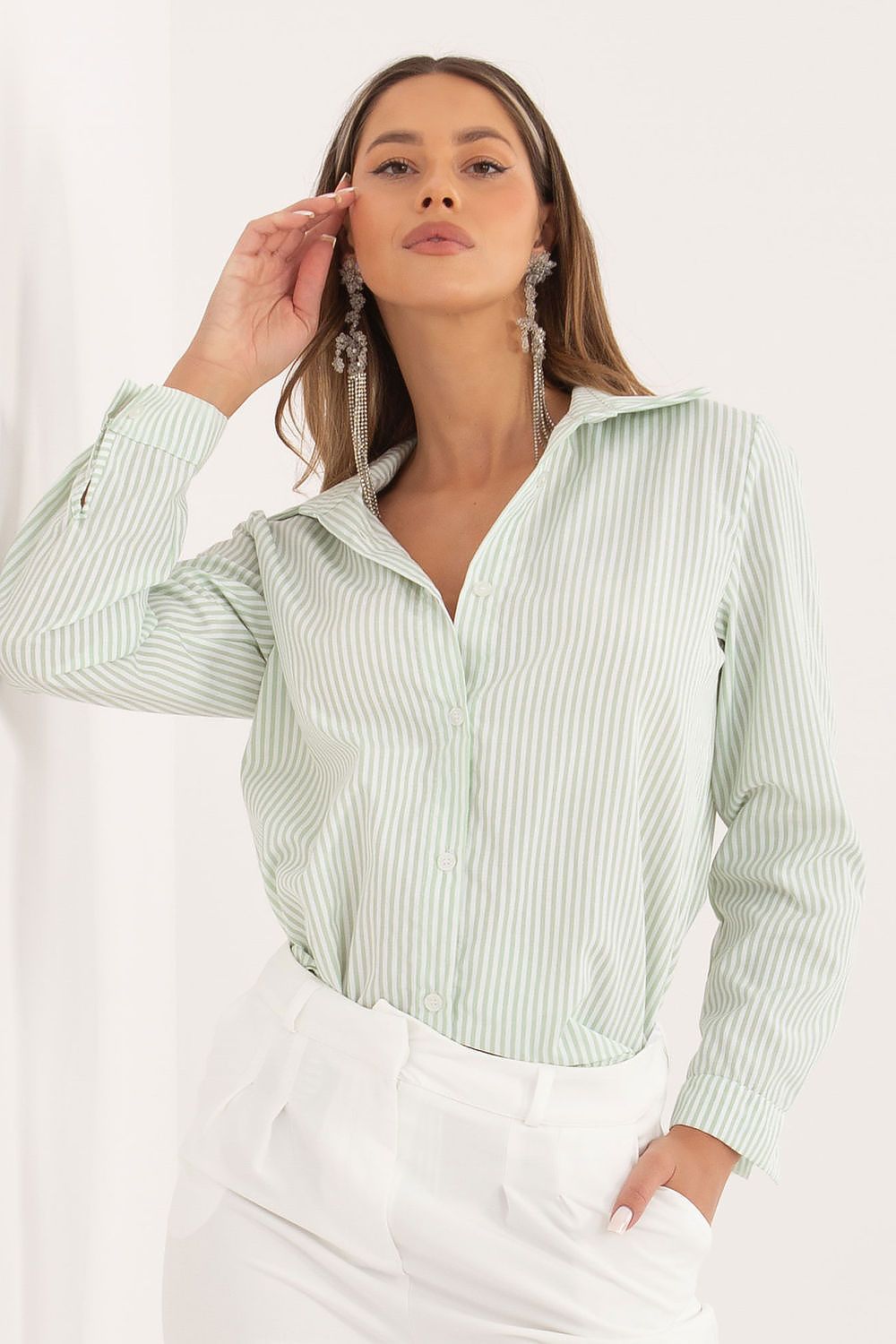 A woman wearing a light sage green and white vertically striped button-up shirt with white trousers, accessorized with long silver dangle earrings, against a clean white background.