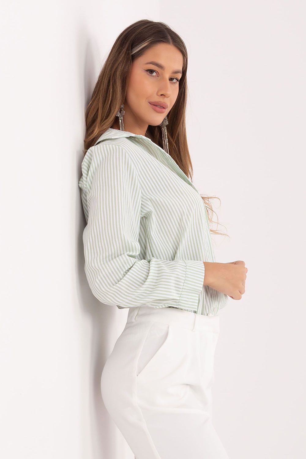 A woman wearing a light sage green and white vertically striped long-sleeve blouse tucked into white pants, posing against a plain white wall.