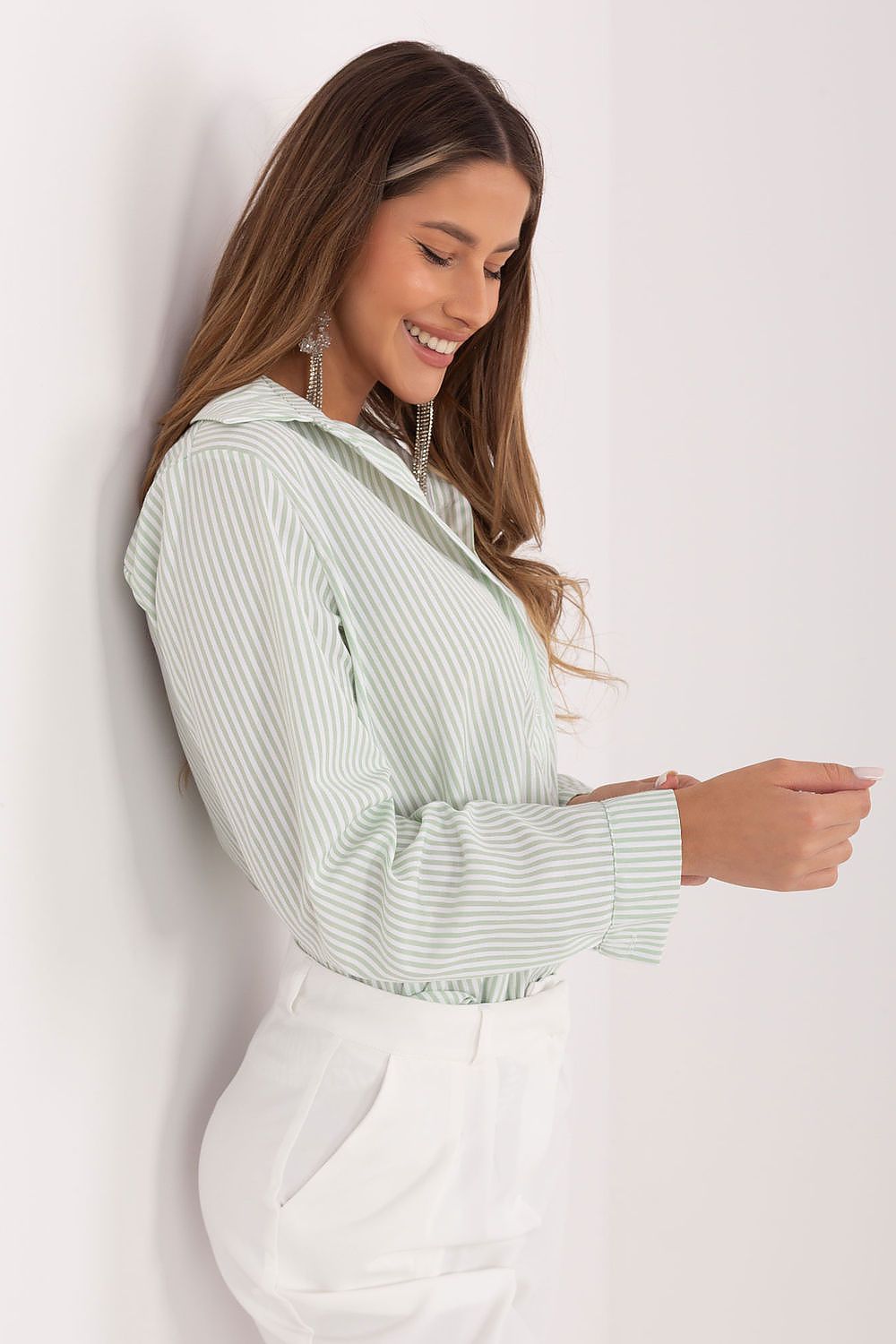 A woman wearing a light green and white vertically striped long-sleeve collared shirt tucked into white pants, leaning against a plain white wall.