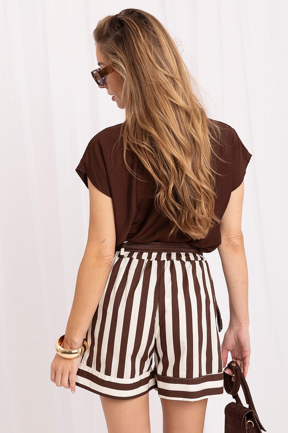 A woman shown from the back wearing a brown short-sleeve top and brown-and-white vertically striped shorts, with sunglasses and a small handbag against a white curtain background.