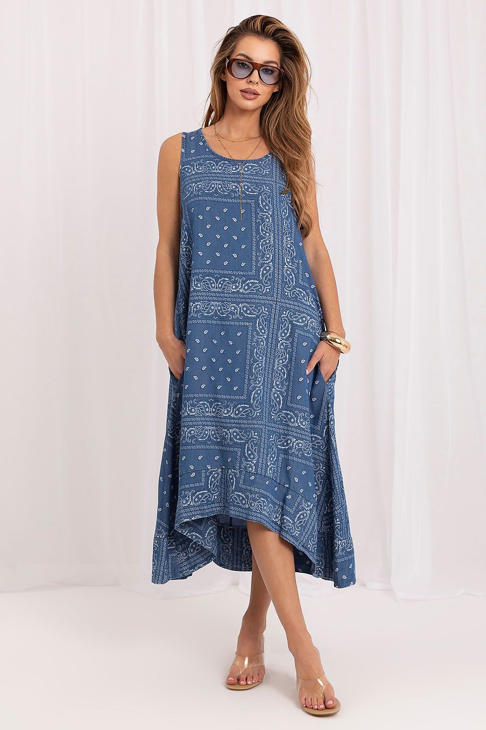 A woman modeling a sleeveless high-low dress with a blue and white bandana print, standing against a white curtain backdrop.