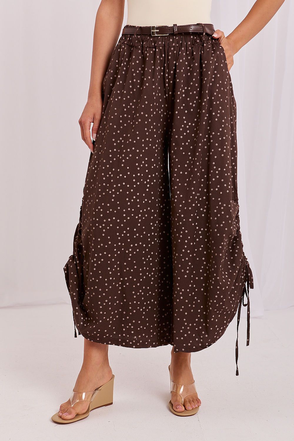 A model wearing brown wide-leg cropped pants with a small white polka dot pattern, side tie details, and a matching brown belt, paired with beige wedge sandals.
