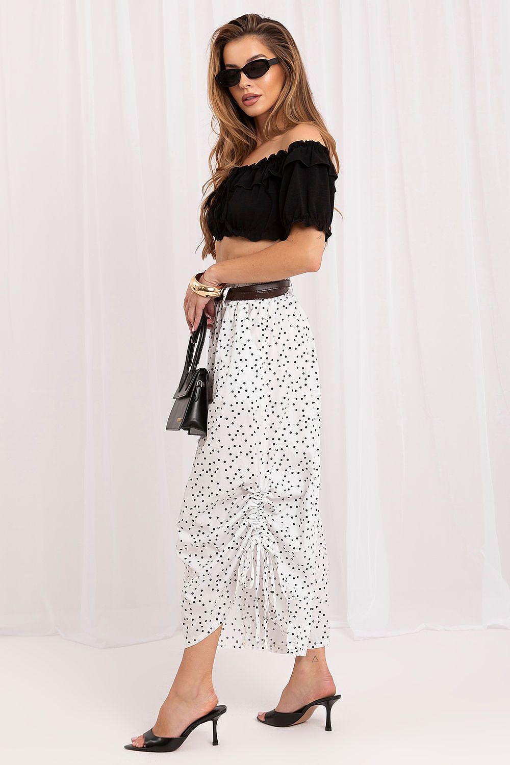 A woman modeling a white midi skirt with a black polka dot pattern, styled with a black off-the-shoulder crop top, a brown belt, black sunglasses, and black mule heels against a white draped backdrop.
