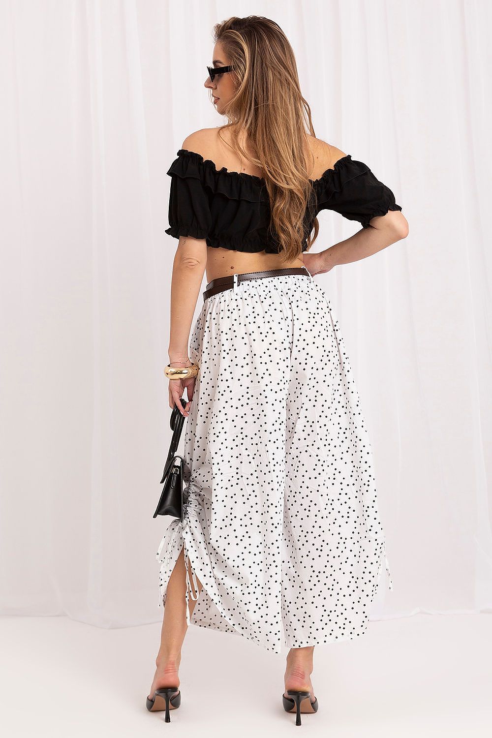 A woman is shown from the back wearing a black off-the-shoulder ruffled crop top and a white maxi skirt with black polka dots. She is holding a small black handbag and wearing black pointed-toe mule heels against a white draped studio backdrop.