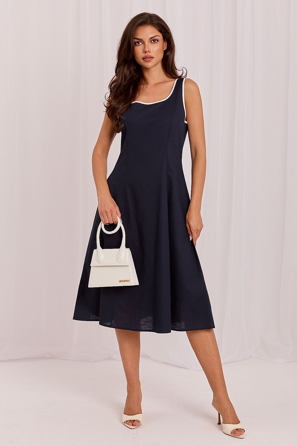 A woman modeling a sleeveless navy midi dress with white trim, holding a small white handbag and wearing white heeled sandals against a light studio backdrop.