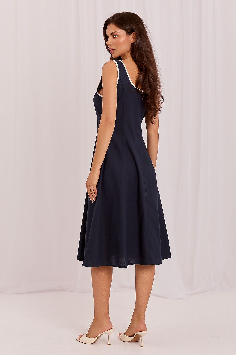 A woman modeling a sleeveless navy blue midi dress with white trim on the straps and armholes, shown from a side-back angle against a white draped studio backdrop.