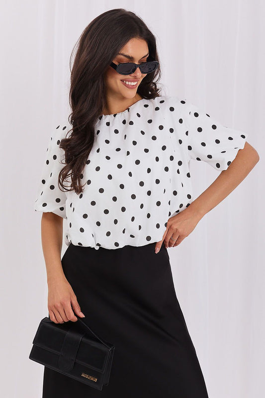 A woman wearing a white short-sleeve blouse with black polka dots and a black skirt, holding a black clutch bag against a light gray background.