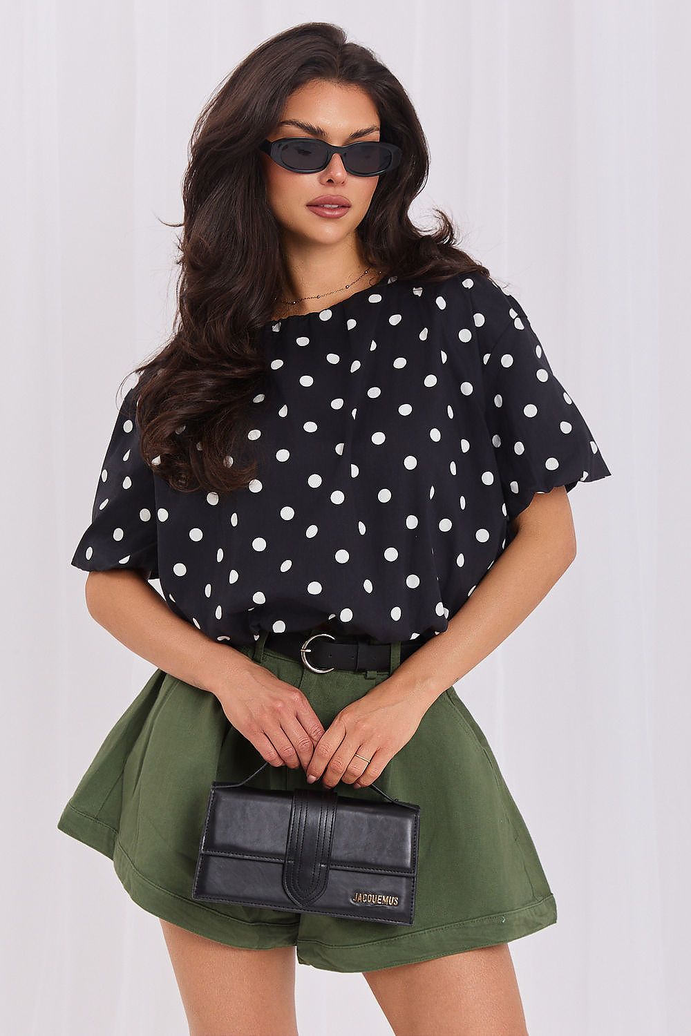 A woman modeling a black short-sleeve top with white polka dots, paired with olive green shorts and a black belt, holding a small black handbag against a light gray studio background.