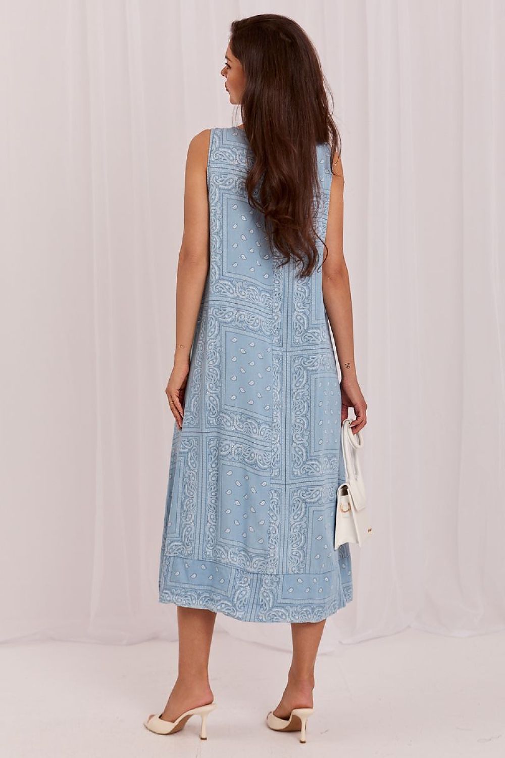 A woman shown from the back wearing a sleeveless light blue midi dress with a white printed border pattern, holding a small white handbag against a white draped backdrop.