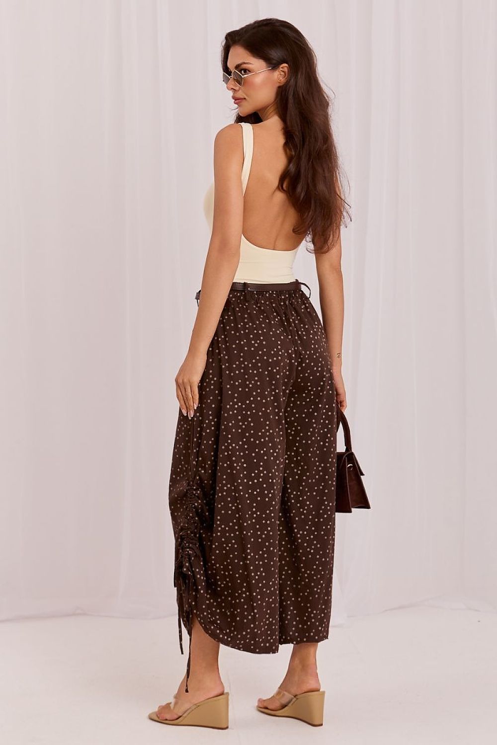 A woman modeling a dark brown wide-leg skirt with a small white polka dot pattern, paired with a cream backless top and beige wedge sandals, shown from a rear three-quarter view against a white draped backdrop.