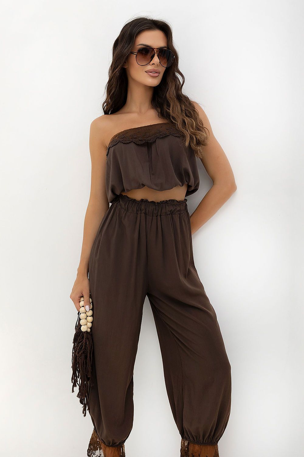 A woman modeling a dark brown two-piece outfit consisting of a ruffled strapless crop top and matching high-waisted loose pants, styled with sunglasses and a beaded clutch against a plain white background.