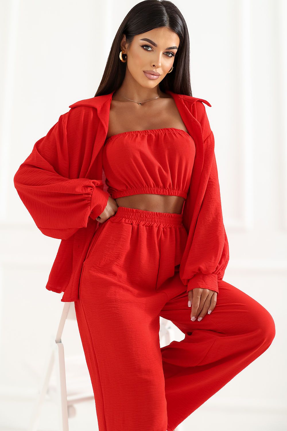 A woman models a bright red two-piece lounge set consisting of a cropped tube top and matching sweatpants, styled with an oversized matching zip-up jacket in a bright white studio setting.