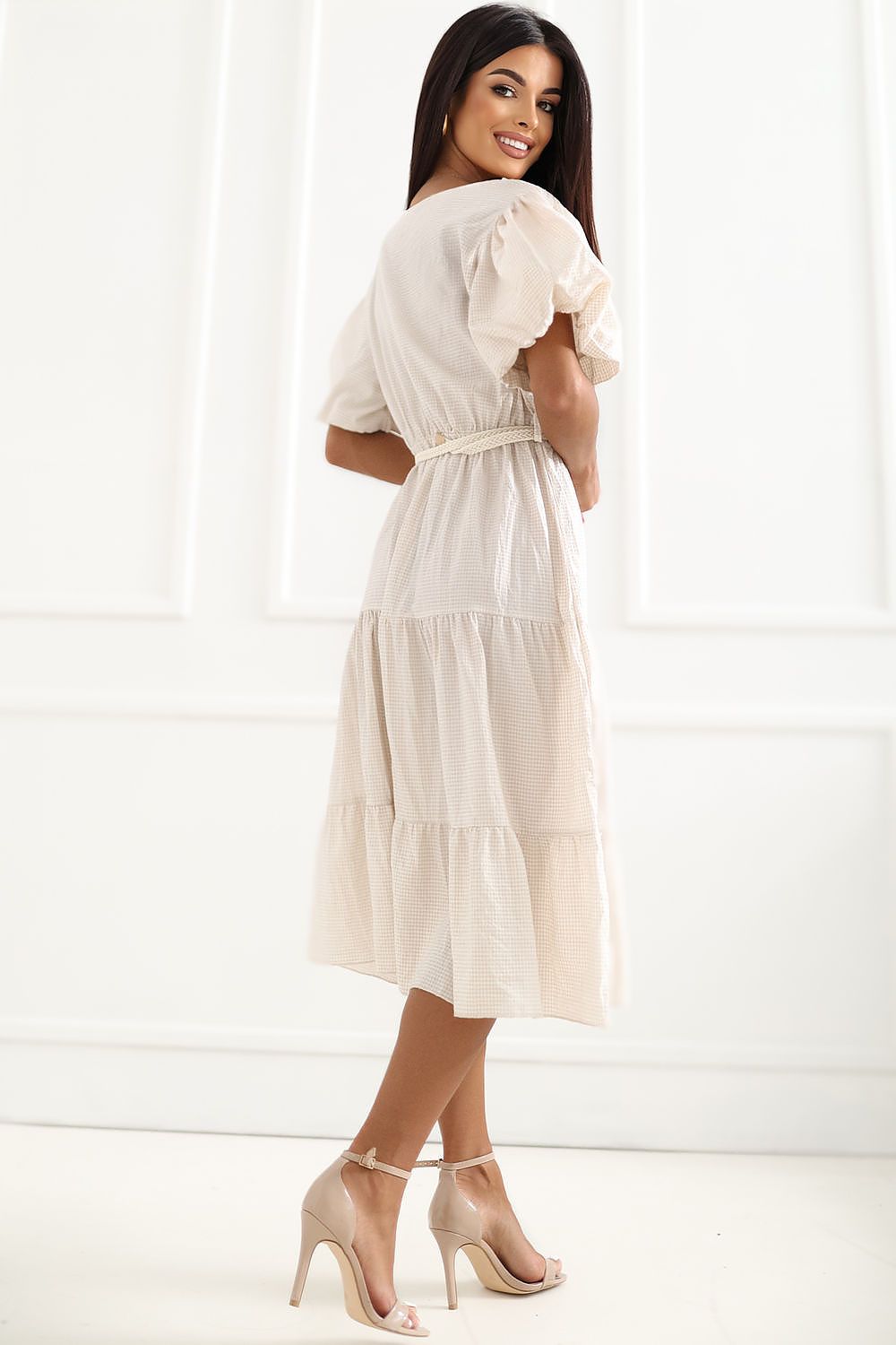 A woman modeling a cream-colored tiered midi dress with puff sleeves and a tie waist, paired with nude high-heeled sandals, set against a white paneled wall.