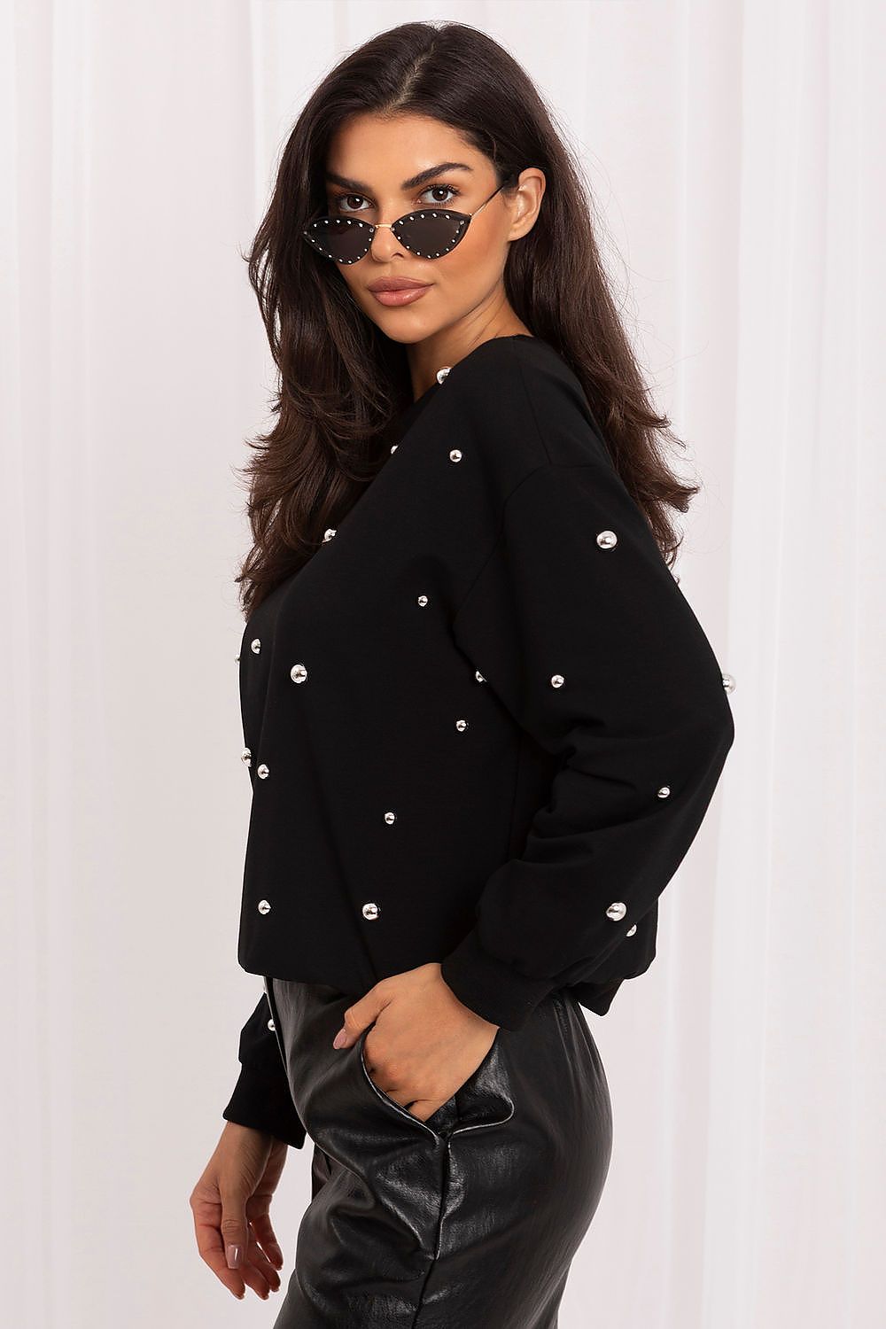 A woman wearing a black long-sleeve knit top with silver rhinestone embellishments, paired with black faux-leather pants, posing against a white curtain background.