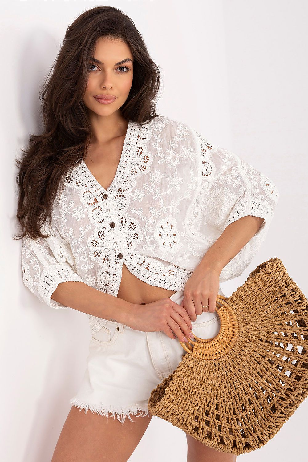 A woman wearing a white lace short-sleeve crop top and white shorts, holding a large woven straw bag against a plain white background.