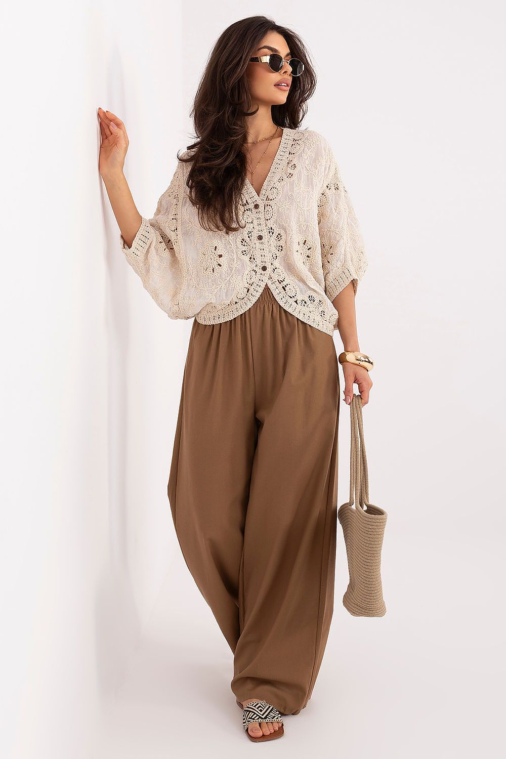 A woman modeling a cream crochet knit cardigan with wide three-quarter sleeves, paired with brown wide-leg pants, holding a beige woven tote bag against a plain white background.