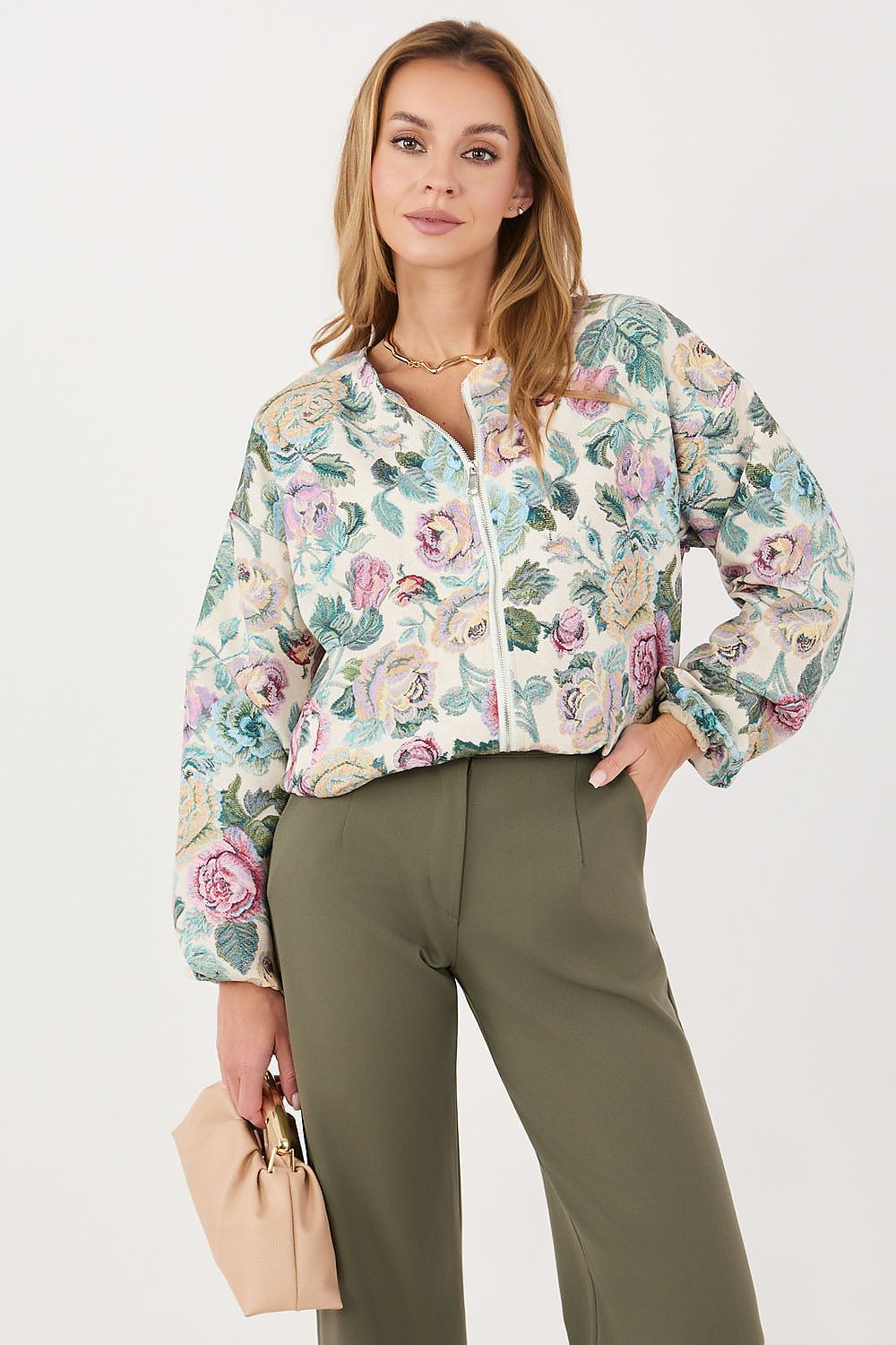 A woman models a light beige floral bomber jacket with green, pink, and blue flowers, paired with olive green trousers and holding a beige handbag against a plain white background.