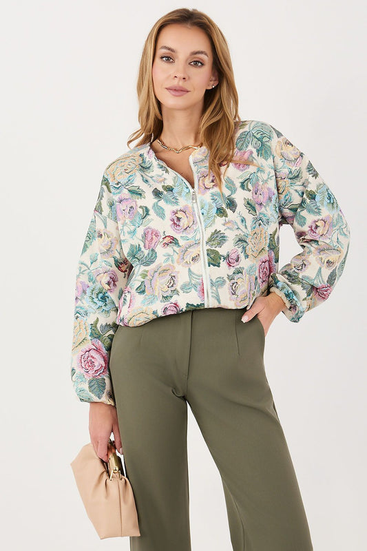 A woman models a light beige floral bomber jacket with green, pink, and blue flowers, paired with olive green trousers and holding a beige handbag against a plain white background.