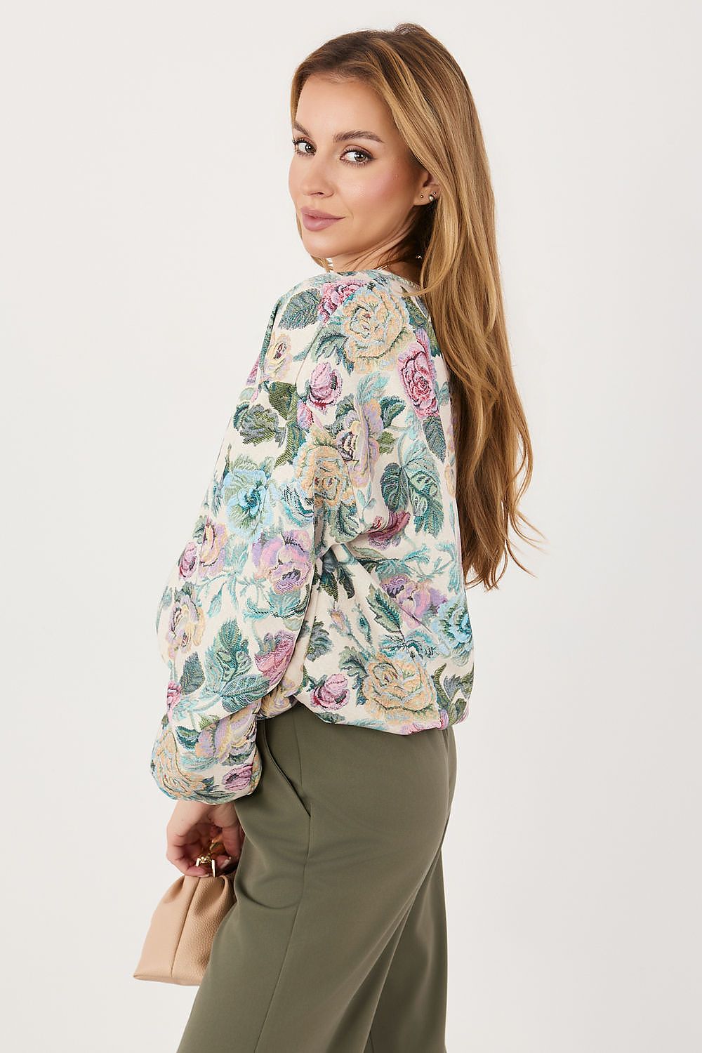 A woman wearing a cream-colored long-sleeve blouse with a colorful floral pattern in pink, green, and blue, paired with olive green pants, holding a small beige clutch bag against a plain light gray background.